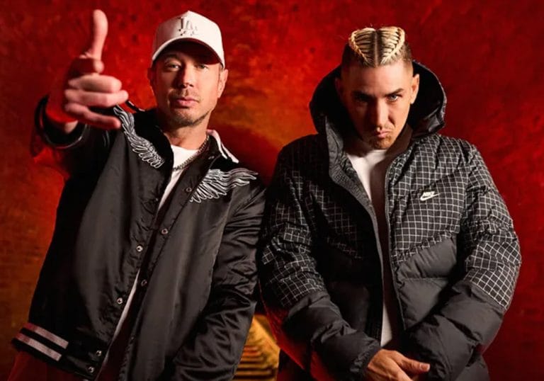Bliss N Eso wearing black puffs jackets, posing in front of a train tunnel that is lit up with red lighting