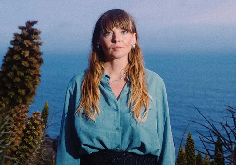 Courtney Marie Andrews portrait standing on the edge of a cliff with bright blue water behind her