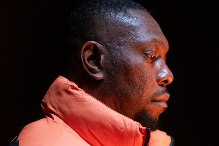 Dizzee Rascal facing side on. Serious still pose, looking downward, wearing a bright orange puffer jacket.