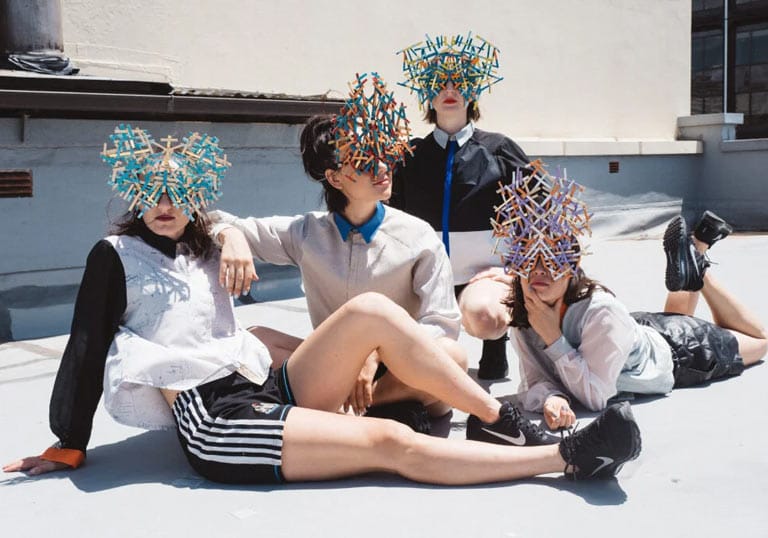 Haiku Hands band, posing on the ground wearing creative masks made of little sticks