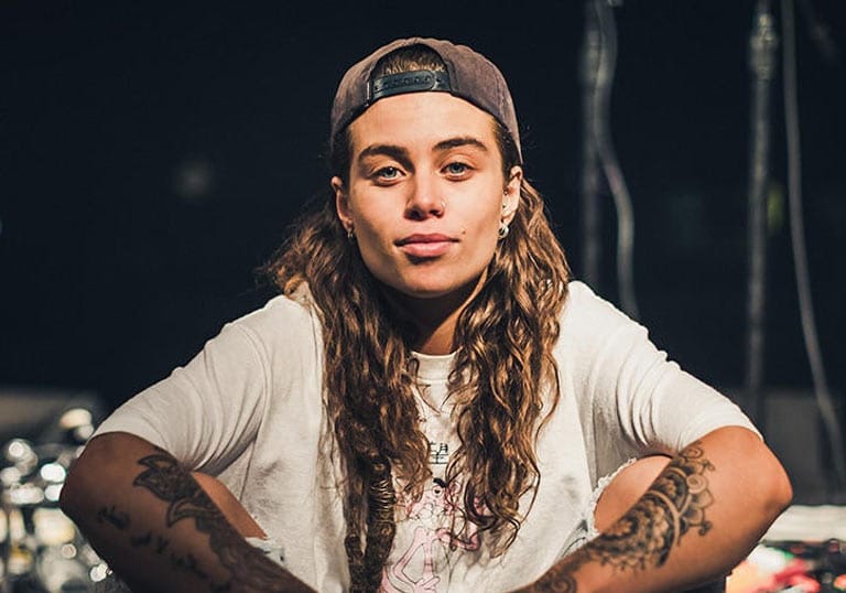 Tash Sultana sitting crossed legged wearing a white T-shirt and brown hat backwards