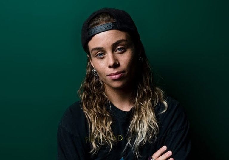 Tash Sultana wearing a hat backwards in front of a dark green wall