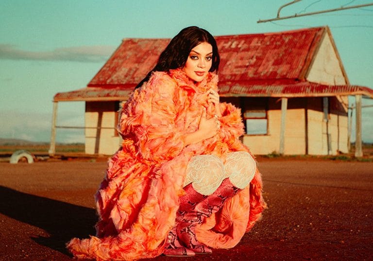 Thelma Plum pictured in a bright ruffly orange coat, standing in a rural area with an old weathered Australian house.