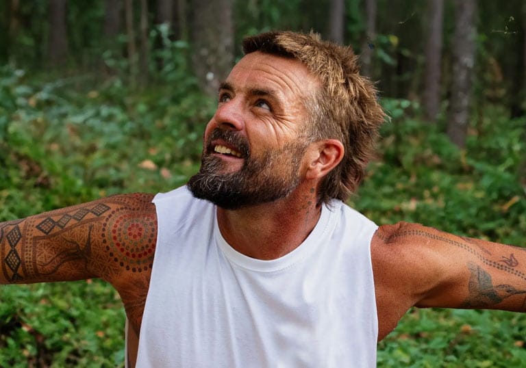 Portrait of Xavier Rudd wearing a white tank top in a forest