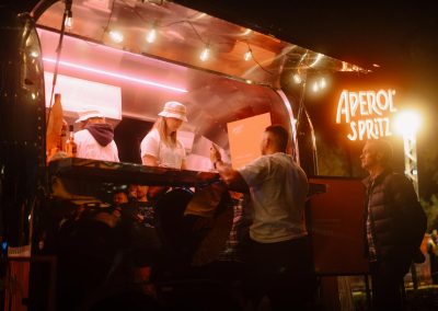 Aperol Spritz Van lit up at night time. Bar staff are dressed in white, serving patrons delicious drinks