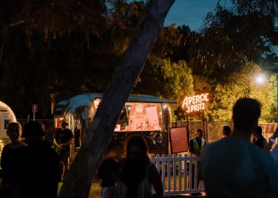 Aperol Spritz Van pictured through the trees at night time with glowing neon sign