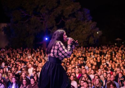 Thelma Plum singing with a microphone. In front of her is a full crowd lit up by the stage lights.